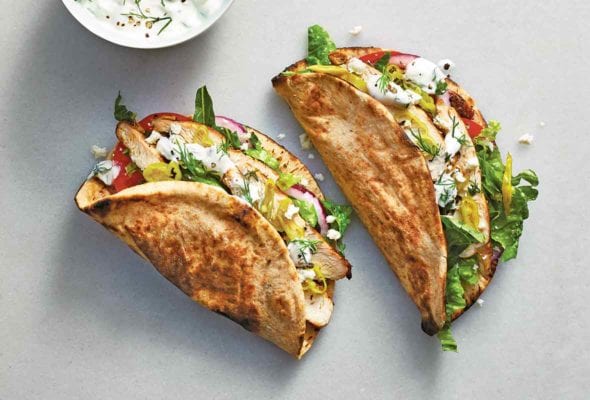Two chicken gyros filled with chicken, red onion, romaine, tomato, and tzatziki with a bowl of tzatziki beside them.
