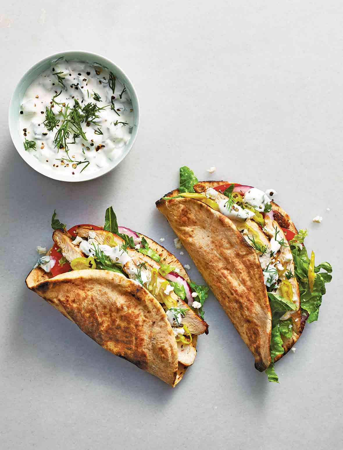 Two chicken gyros filled with chicken, red onion, romaine, tomato, and tzatziki with a bowl of tzatziki beside them.