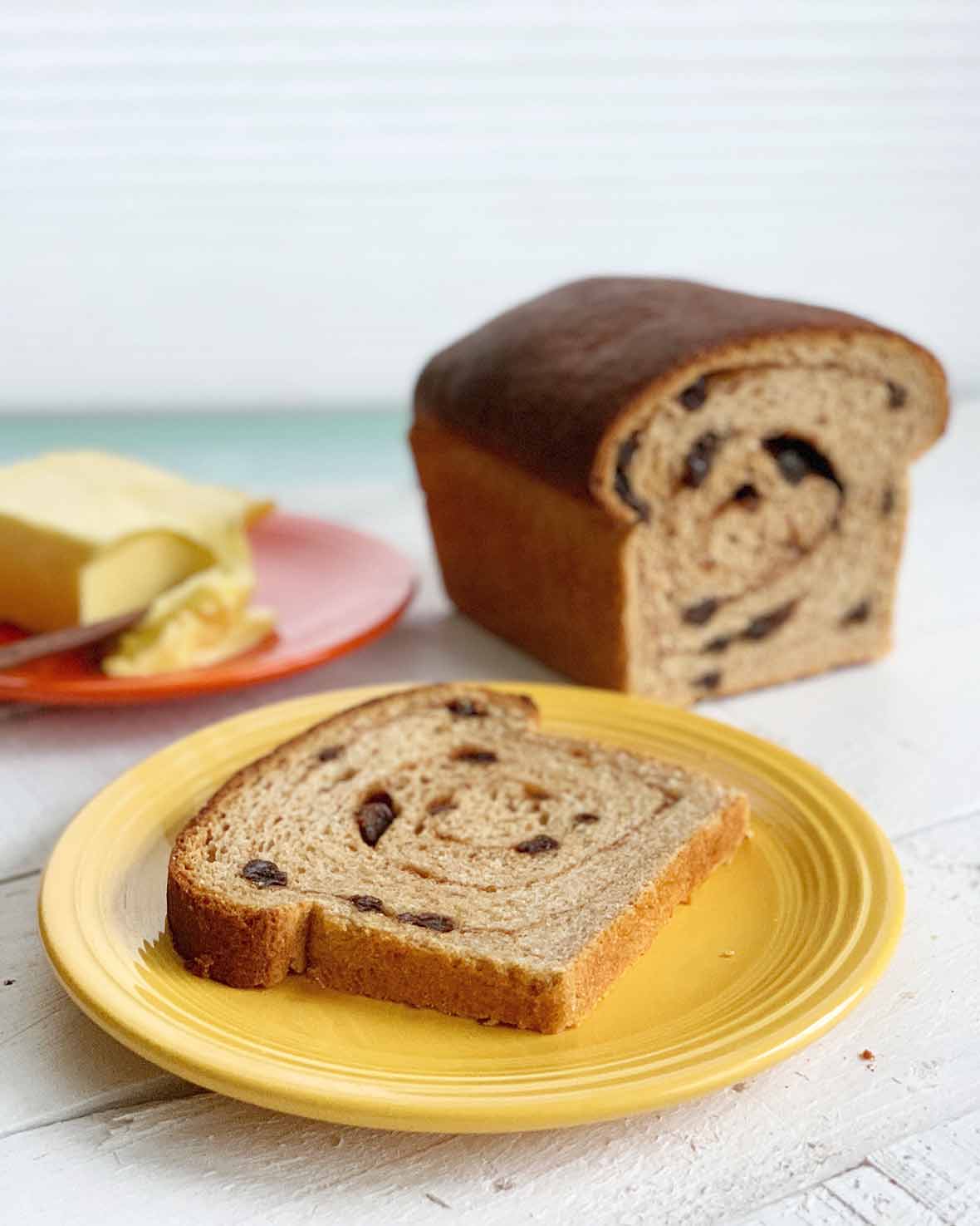 A cut loaf of cinnamon raisin swirl bread and a slice of it on a yellow plate with butter in the background