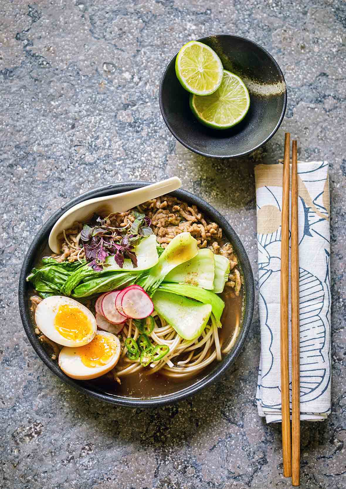 A black bowl filled with curried pork ramen, jammy egg, bok choy, sliced radish, and basil.