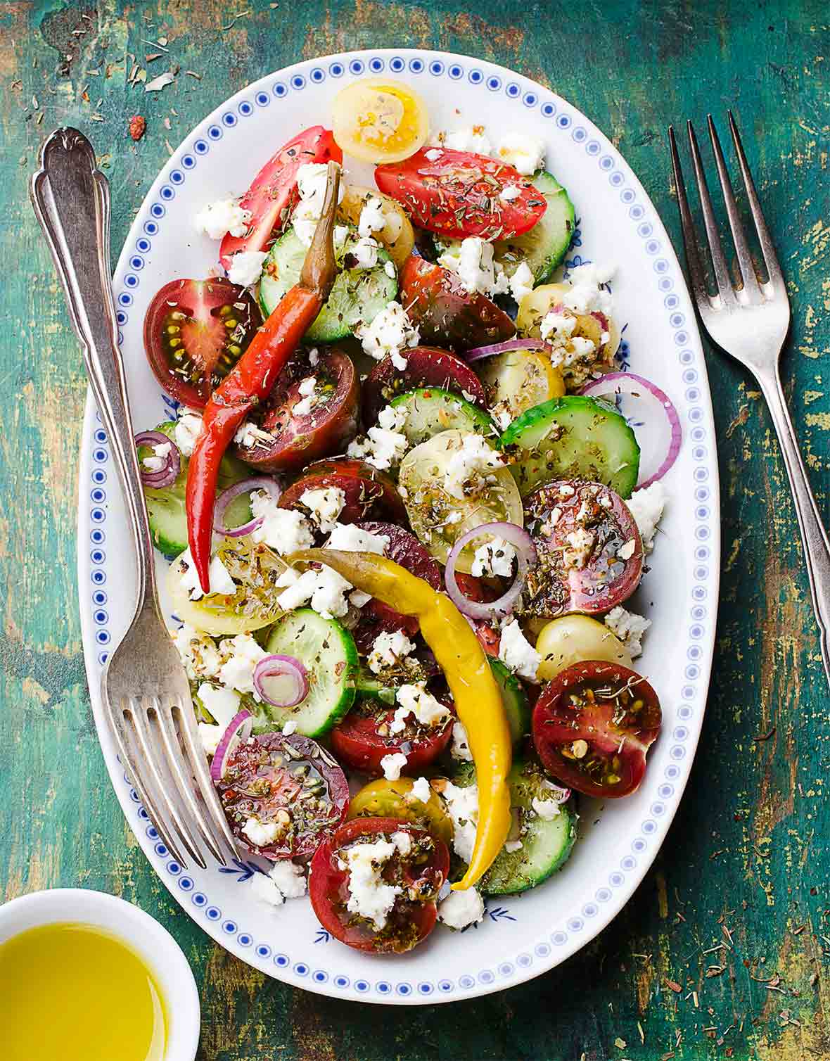 A white oval platter filled with Greek salad, with tomatoes, onions, cucumbers, peppers, and feta.