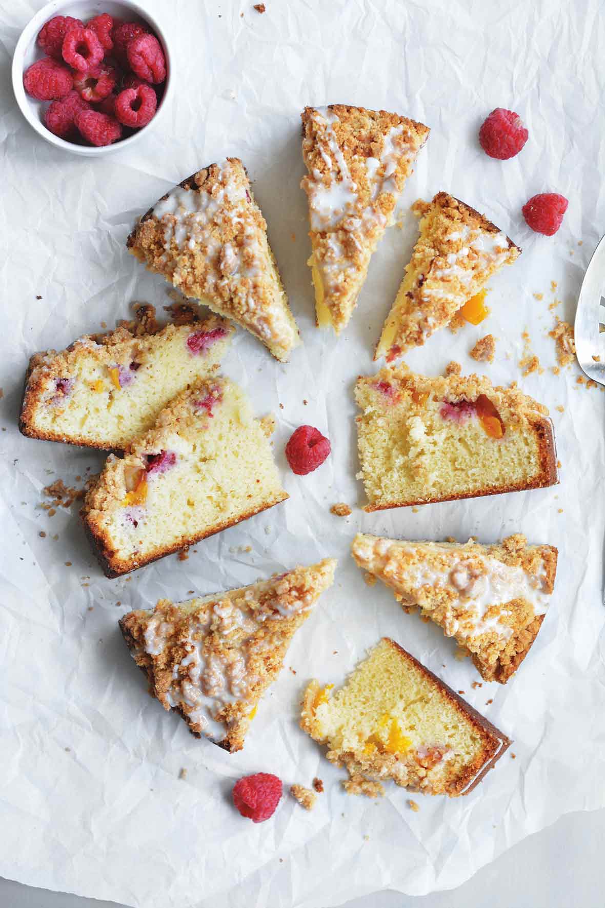 Nine slices of raspberry peach coffee cake with a bowl of raspberries nearby and some more raspberries scattered around the cake.