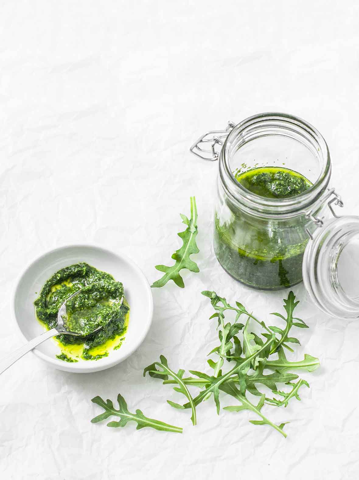 A white bowl partially filled with arugula pesto and a spoon resting in the pesto. A few arugula leaves scattered beside the bowl.