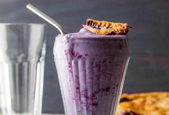 A tall glass filled with blueberry pie milkshake, topped with a piece of pie crust.