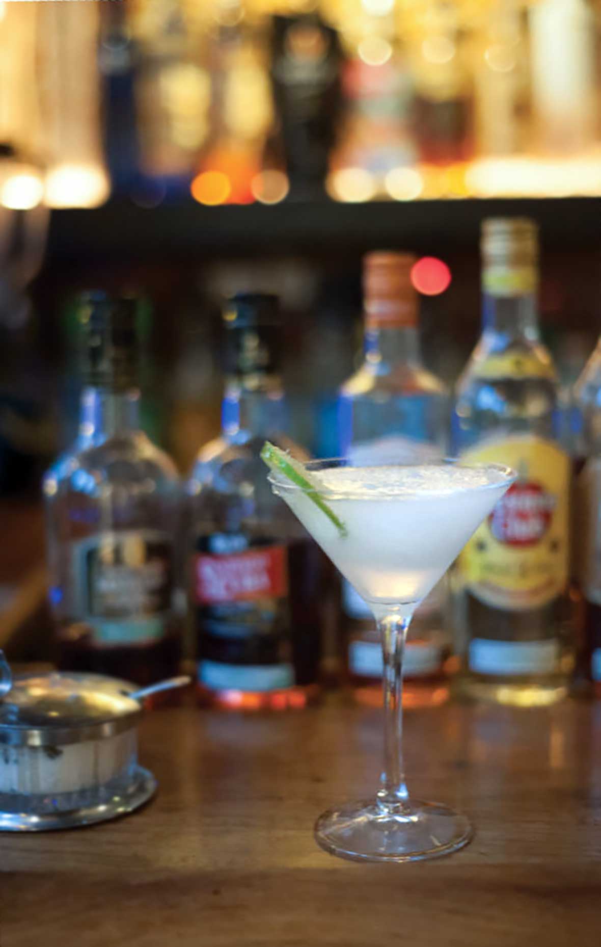 A martini glass filled with caipirinha and a lime wheel on a bar with liquor bottles in the background.