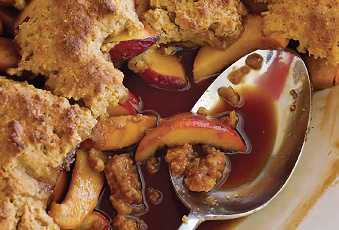 A spoon resting in a dish of cornmeal drop-biscuit peach cobbler.