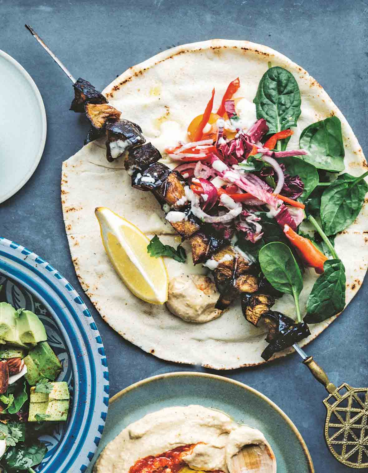 An eggplant kebab on a pita with a lemon wedge, garlic sauce, spinach leaves, red onion, cherry tomatoes, red pepper, and cabbage.