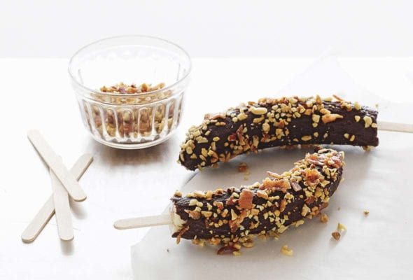 Two frozen chocolate bacon bananas on sticks with a dish of crushed peanuts and extra popsicles sticks beside them.
