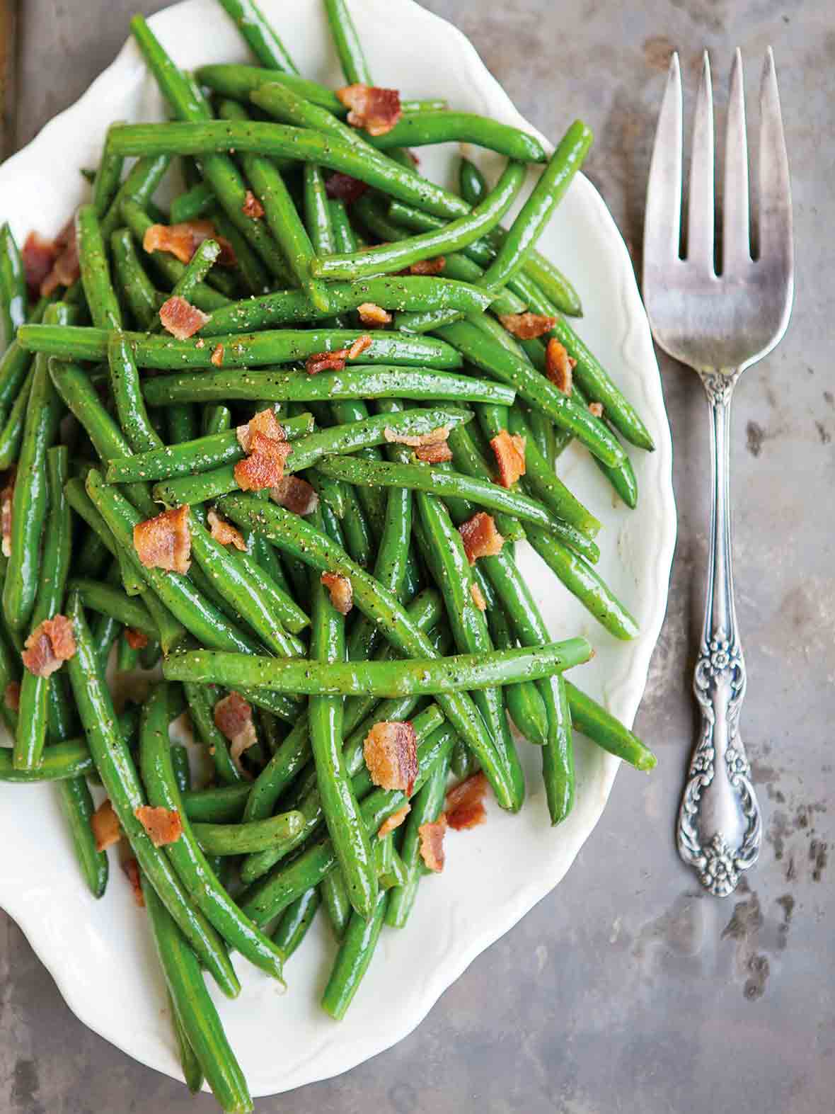 A white serving platter filled with sweet and sour green beans, topped with crumbled bacon.