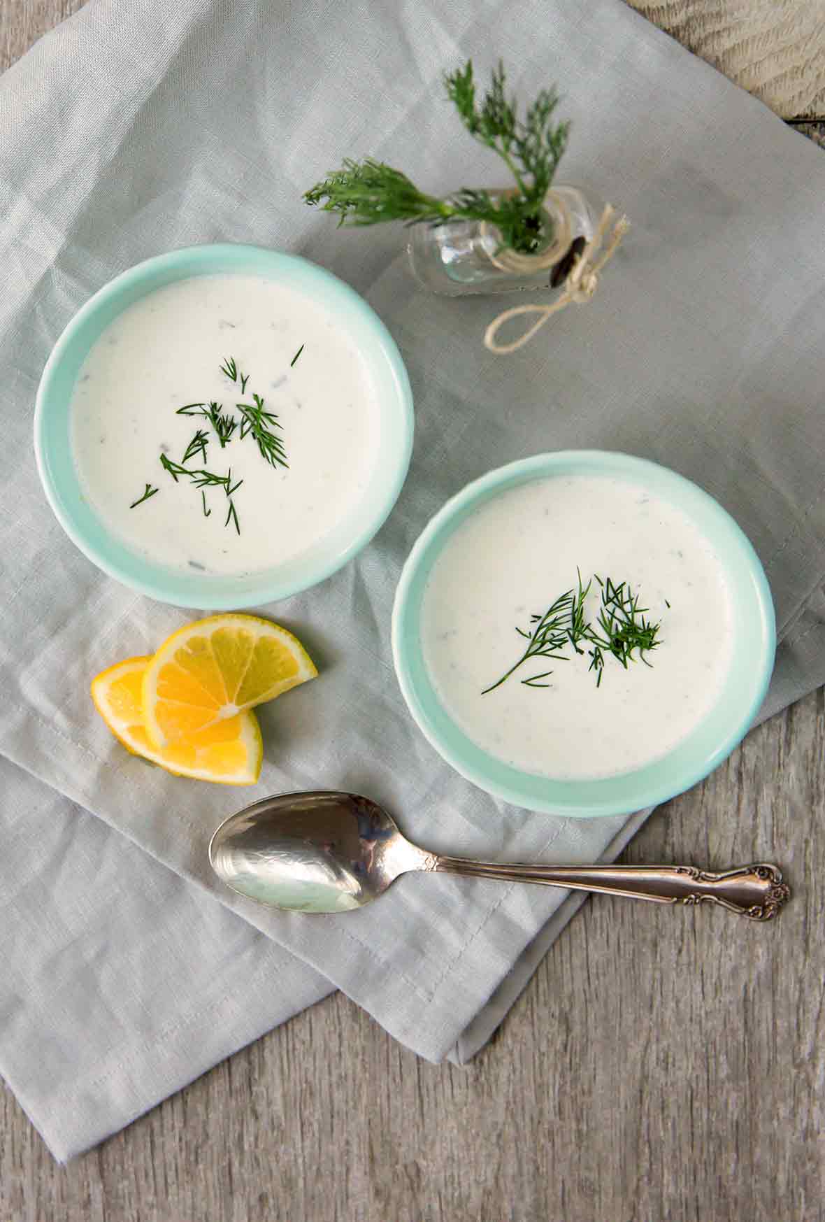 Two blue bowls of cold cucumber soup with yogurt and dill on a linen cloth with a spoon and lemon beside them.