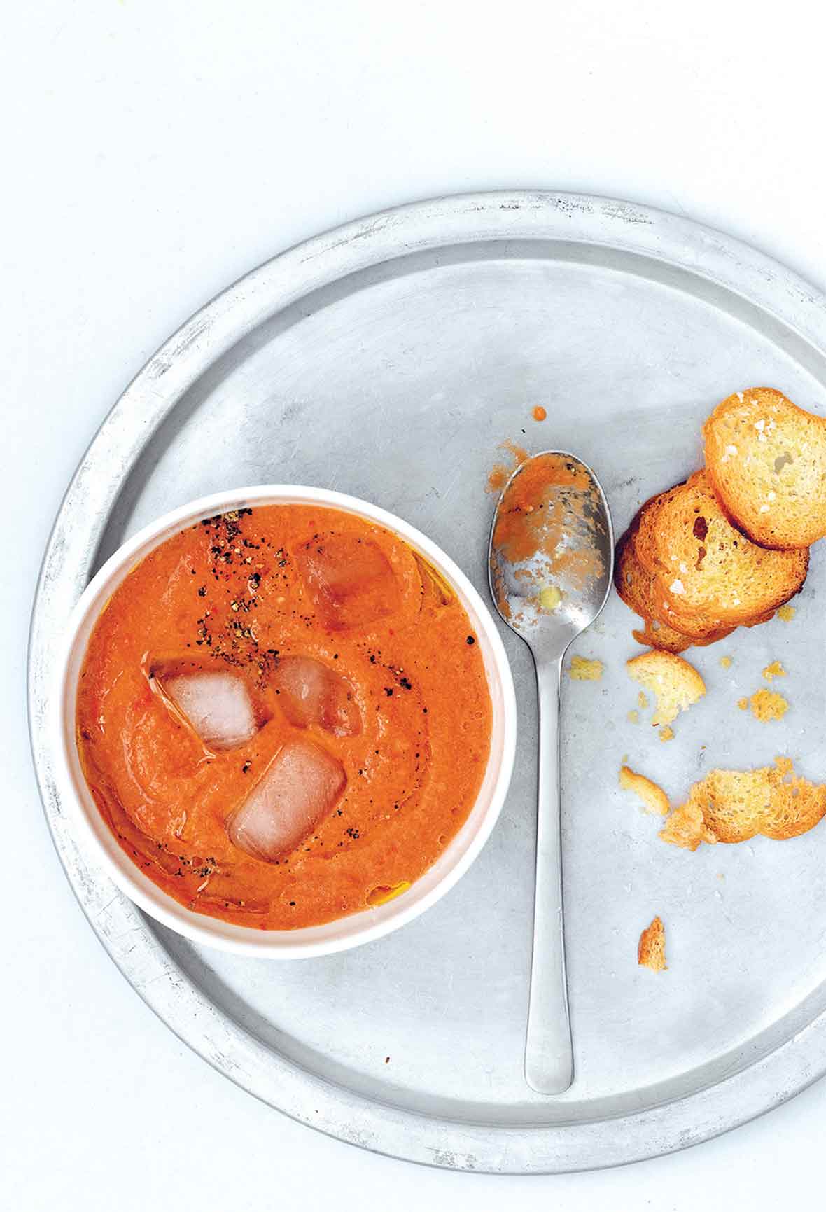 A bowl of easy gazpacho with ice cubes floating in it on a silver plate with a spoon and a few crostini.