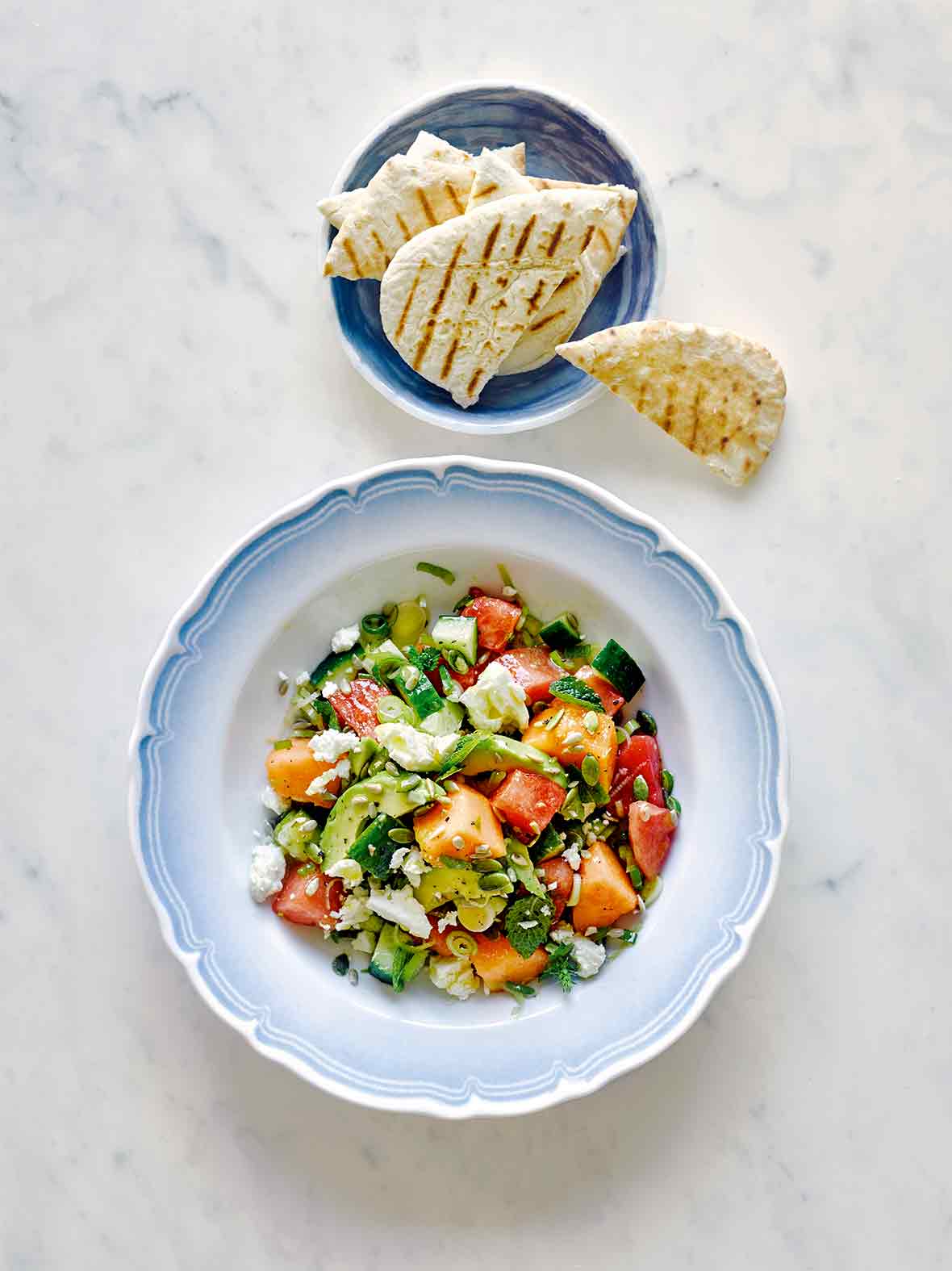 A blue and white bowl filled with melon and avocado salad with a bowl of pita beside it.