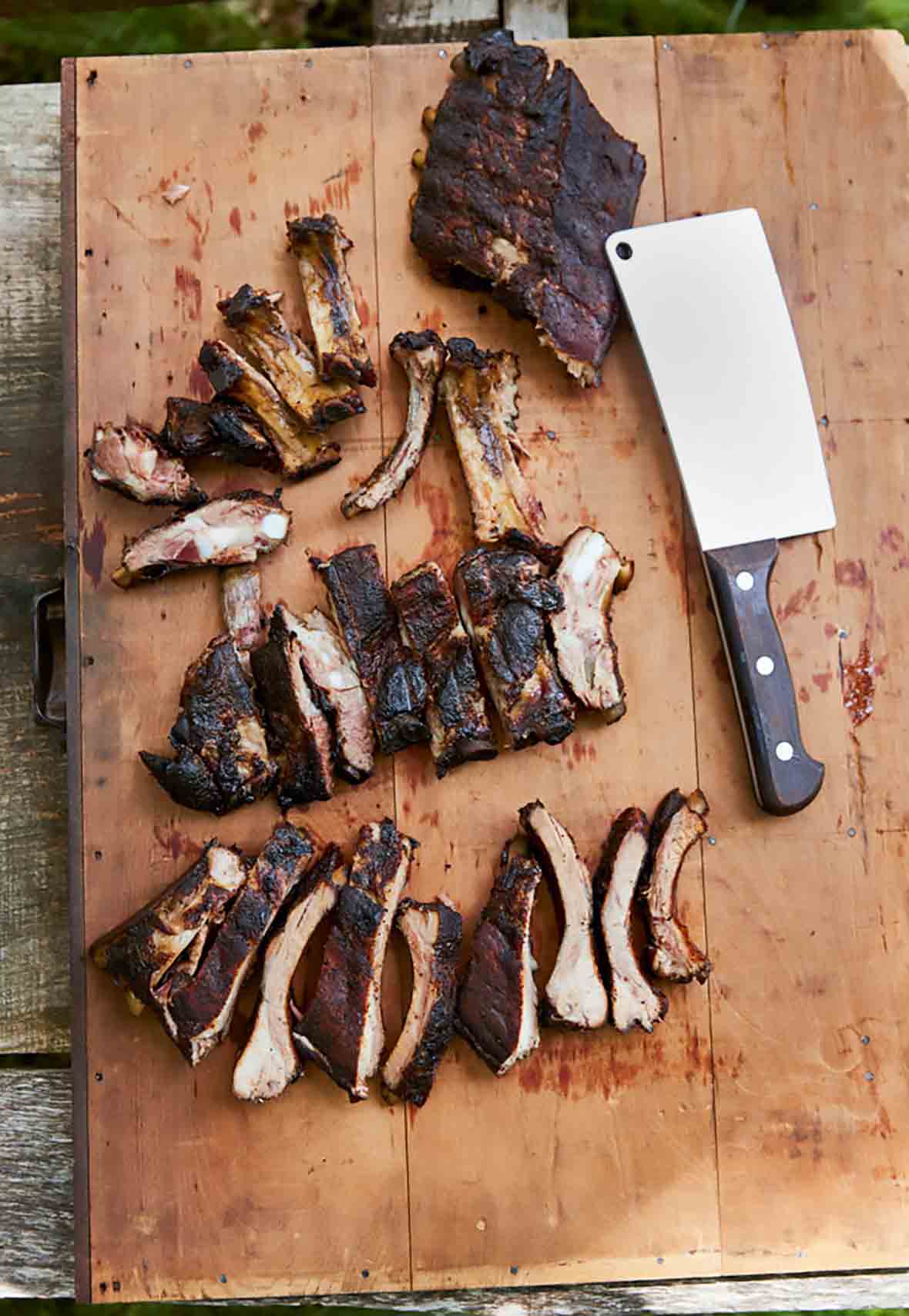 A wooden cutting board topped with a rack of smoked pork ribs, some cut into individual ribs, and a cleaver lying beside them.