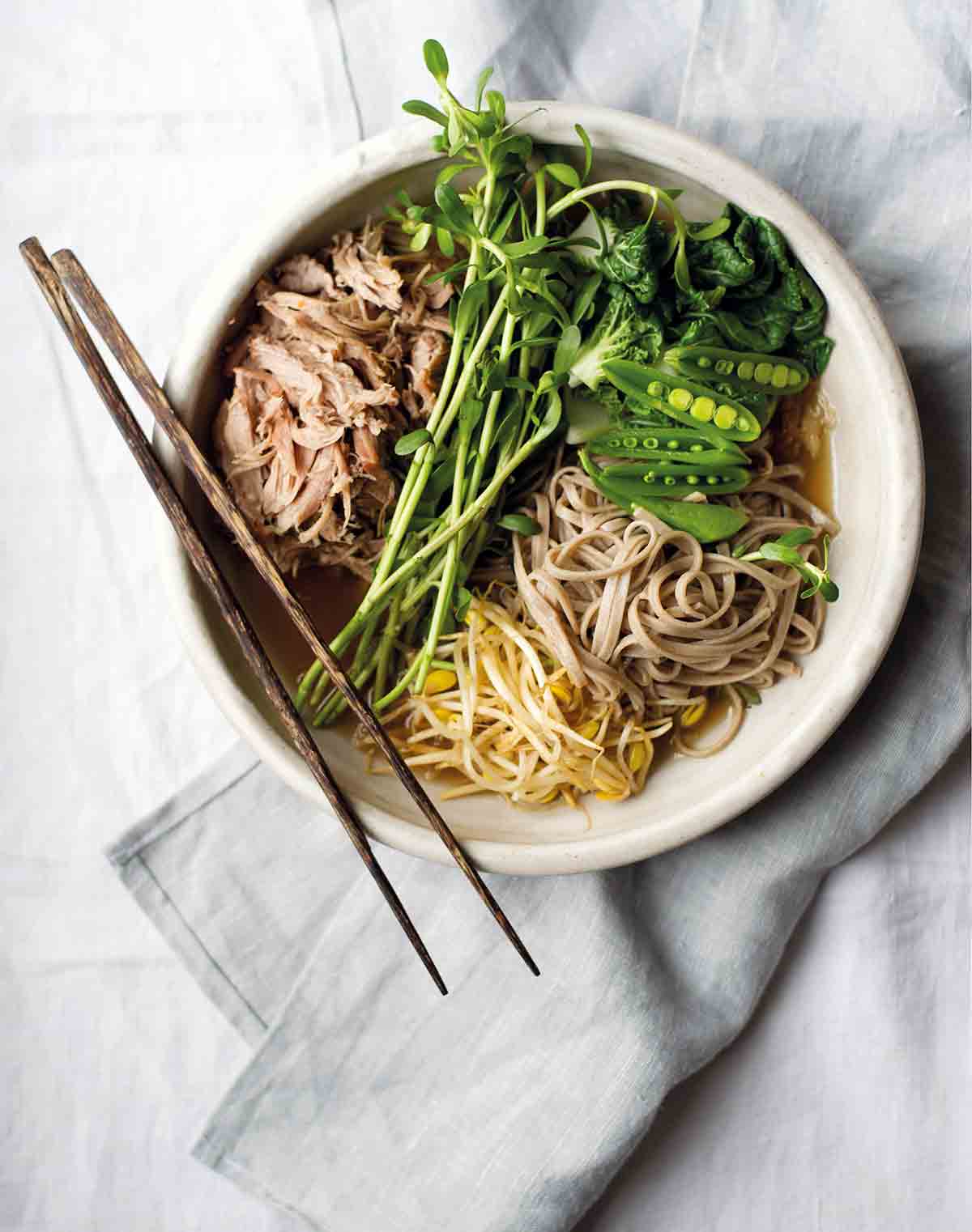 A bowl of chashu pork ramen, filled with braised pork, soba noodles, bean sprouts, peas, and bok choy, with a pair of chopsticks resting on top.
