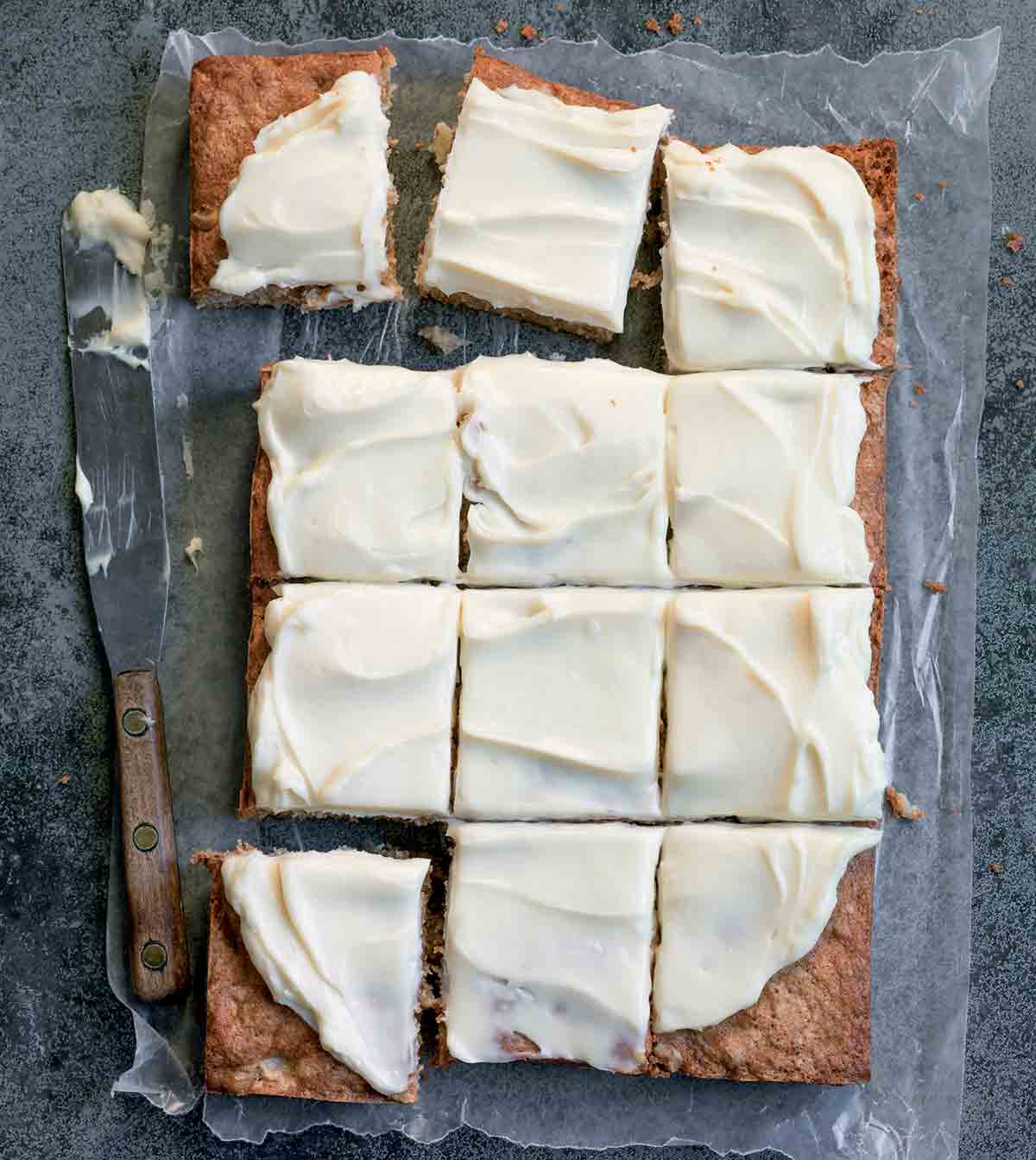 A frosted apple cake on a sheet of wax paper that has been cut into 12 squares with a knife resting beside.