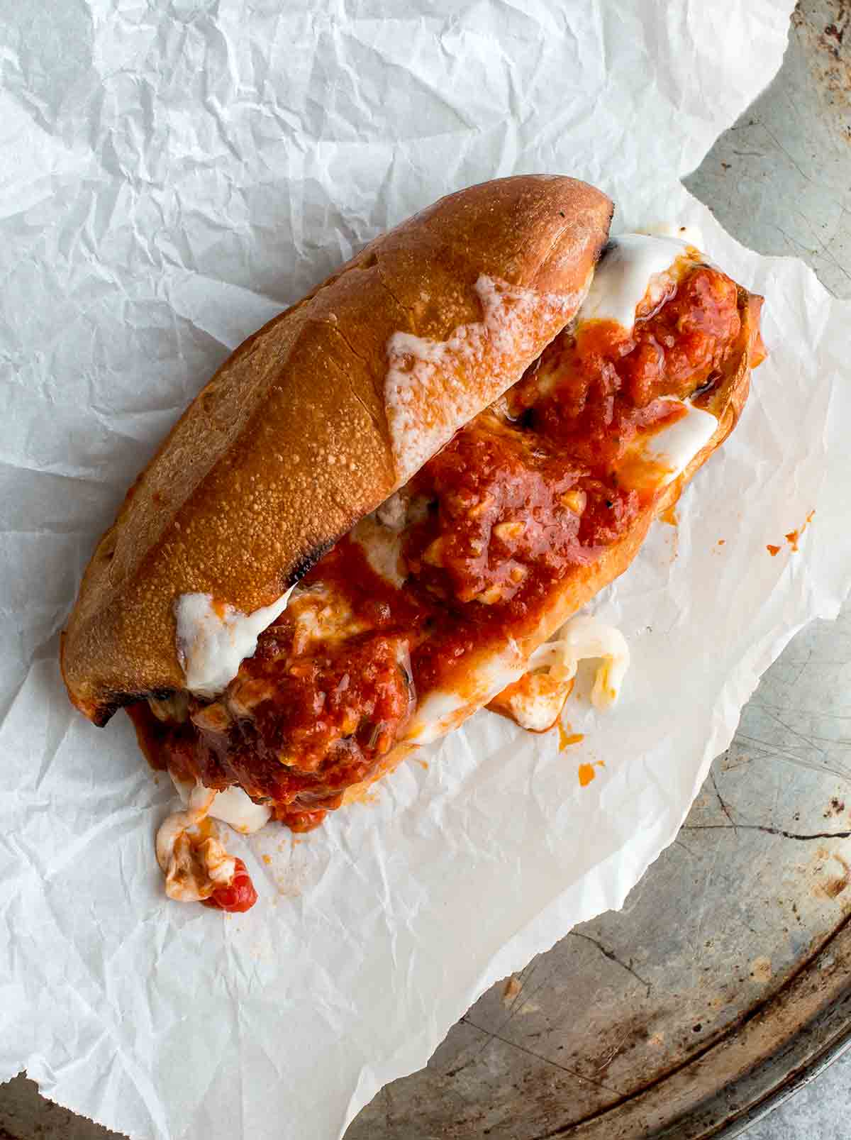 A harissa meatballs sandwich, made with three meatballs stuffed in a sub roll along with cheese and topped with tomato sauce on a sheet of parchment paper.