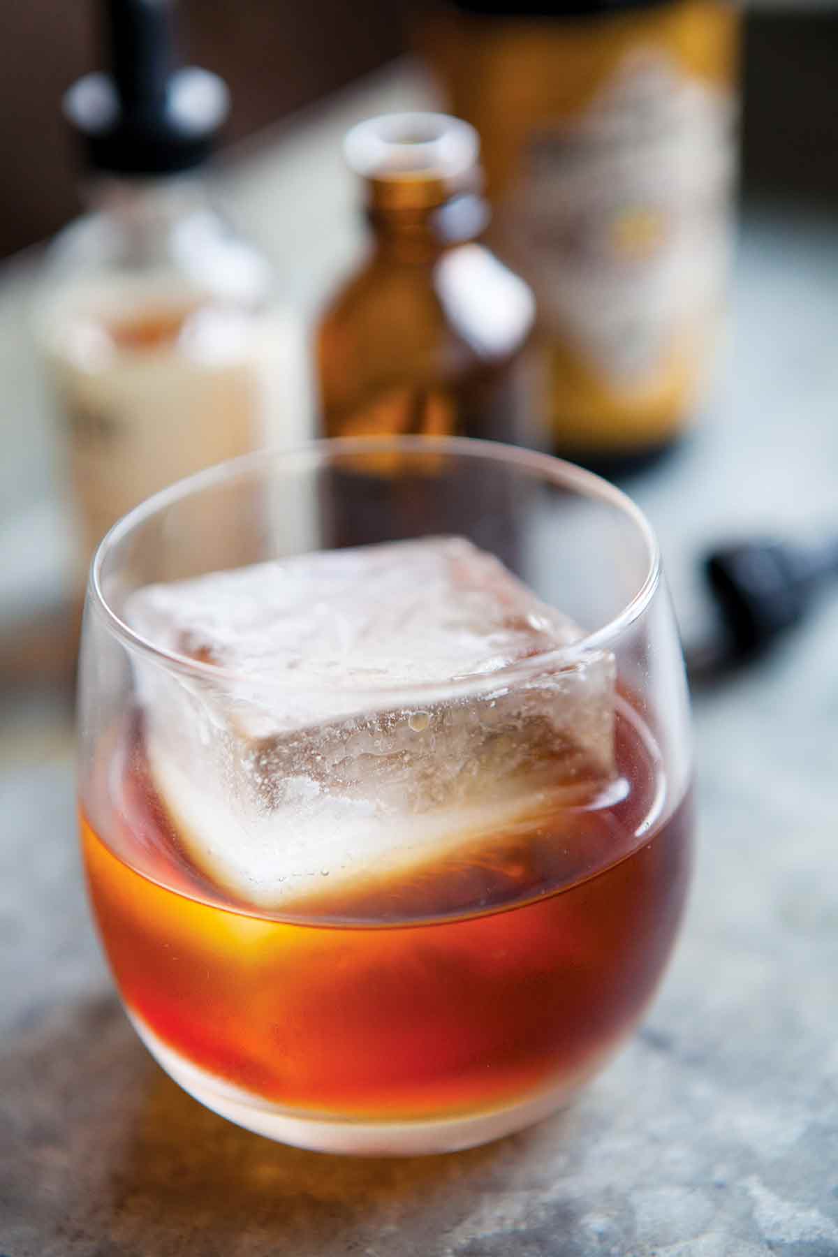 A high ball glass partially filled with an amber Sazerac cocktail without absinthe and an oversized ice cube.