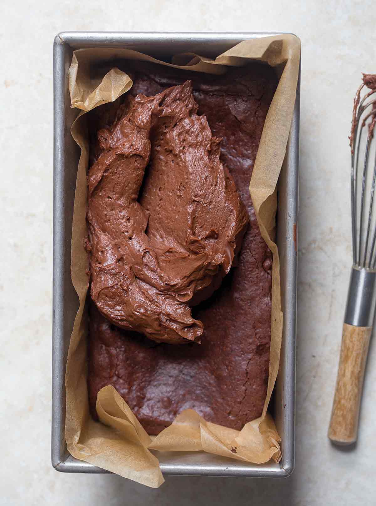 A parchment lined loaf pan filled with partially frosted small batch brownies and a wire whisk on the side.