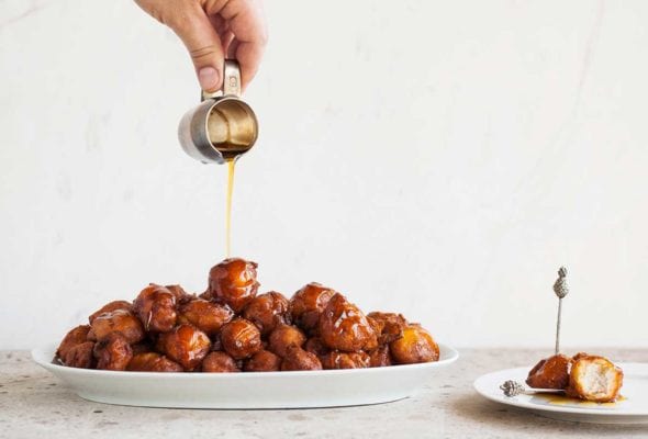 A person pouring spicy maple bourbon syrup over a platter of waffle fried chicken bites, with a white plate with two bites on it beside the platter.