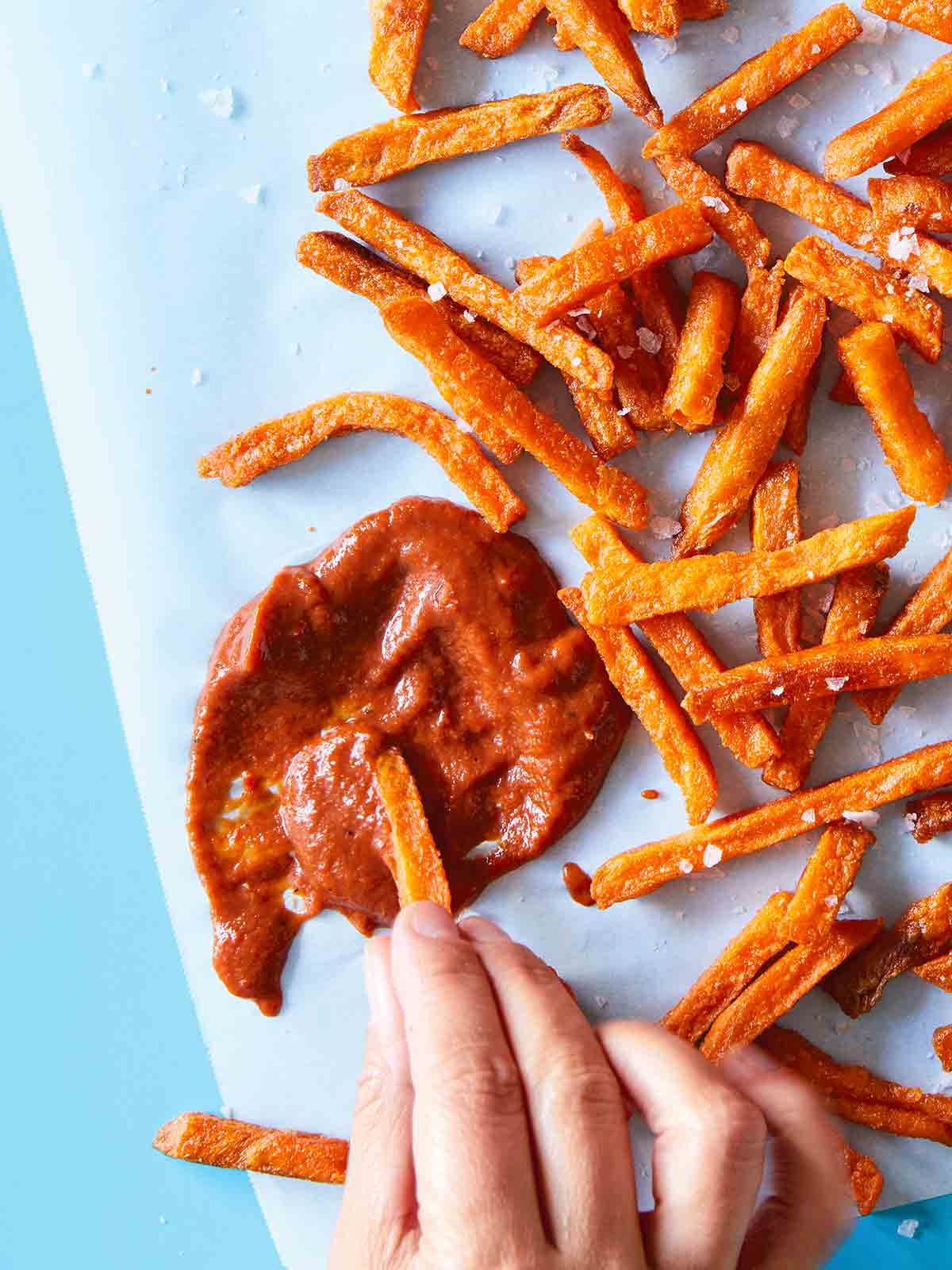 A hand dipping fries into a splatter of bomb hot sauce with a pile of fries scattered beside the sauce.