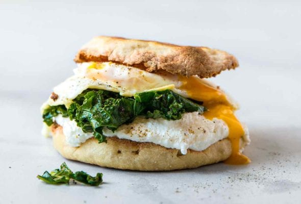 A breakfast sandwich with fried egg, kale, and ricotta sandwiched between two toasted English muffin halves.