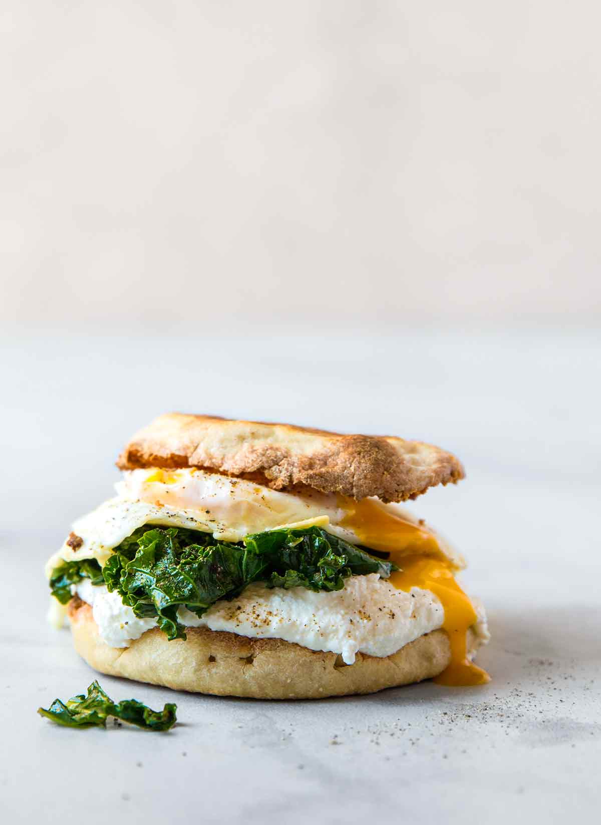 A breakfast sandwich with fried egg, kale, and ricotta sandwiched between two toasted English muffin halves.