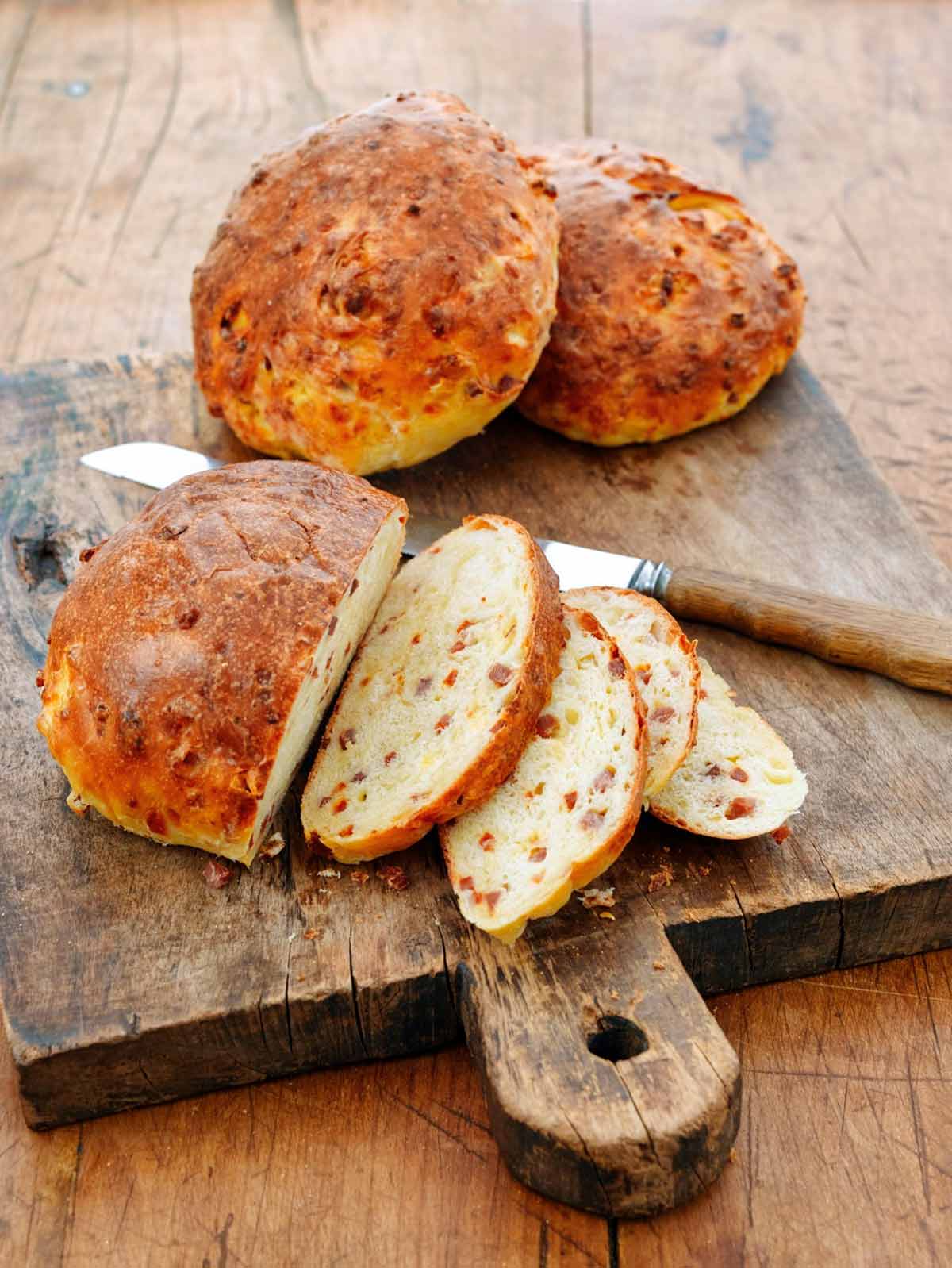Three presunto and cheese loaves on a wooden cutting board, with one cut into several slices and knife lying beside it.