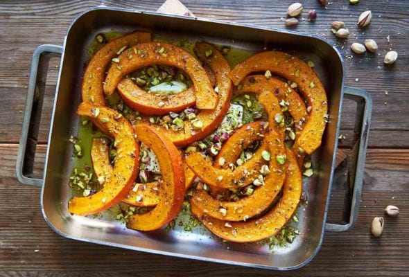 A metal roasting pan filled with roast butternut squash, feta, and pistachios on a wooden table with pistachios scattered around it.