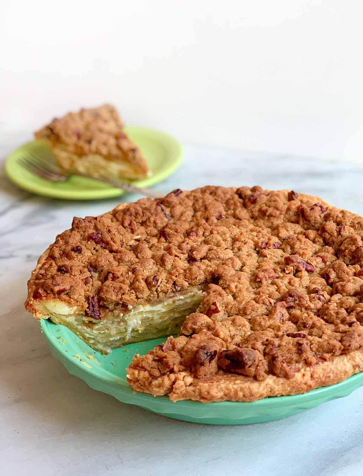 A sour cream apple pie with one slice cut out in a ceramic teal deep pie dish. The slice is on a green plate in the background.