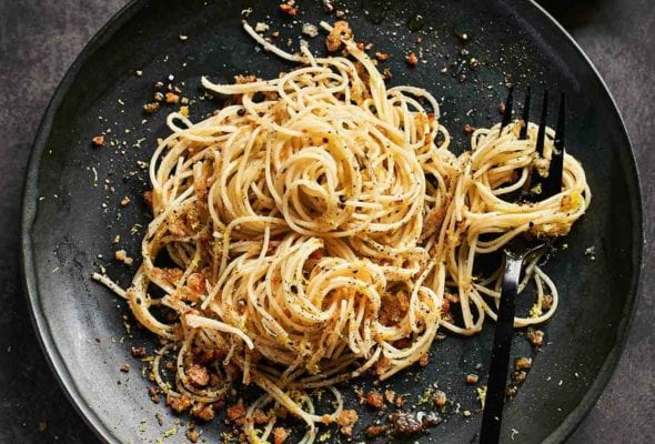 A black plate topped with angel hair pasta with lemon bread crumbs and a fork and a squeezed half lemon rests beside the plate.