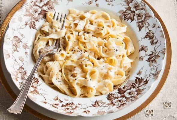 A patterned bowl filled with lemony fettuccine alfredo on a white and gold plate with a glass of water and dish of Parmesan on the side.