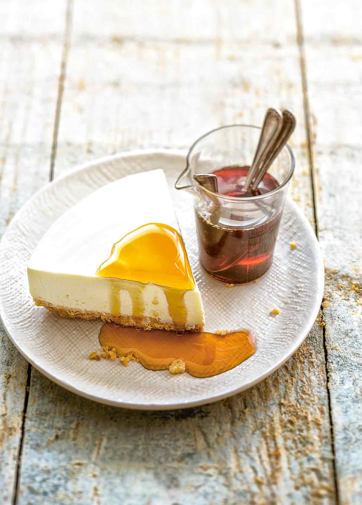 A slice of no-bak cheesecake drizzled with maple bourbon syrup on a white plate with a pitcher of syrup also on the plate.