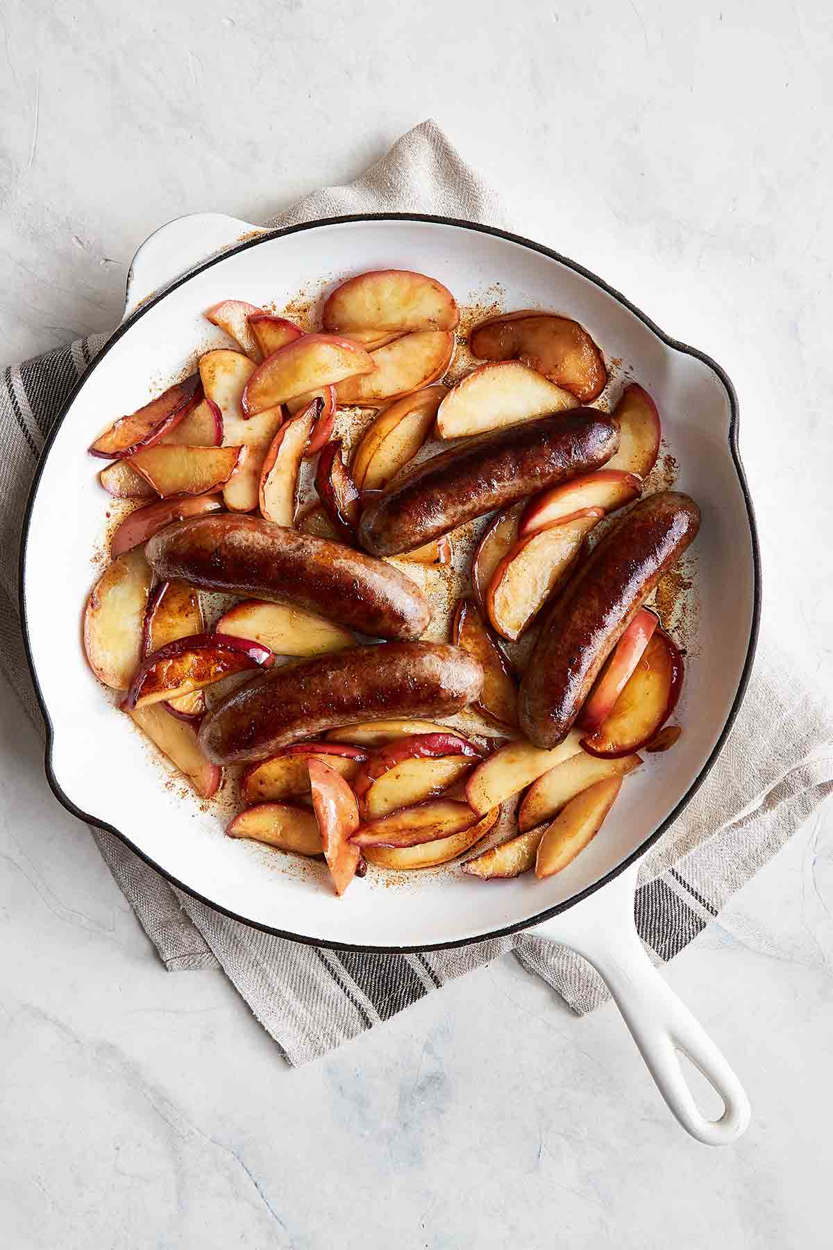 A white enamel cast-iron skillet filled with sautéed sausages with apples on a grey linen cloth.