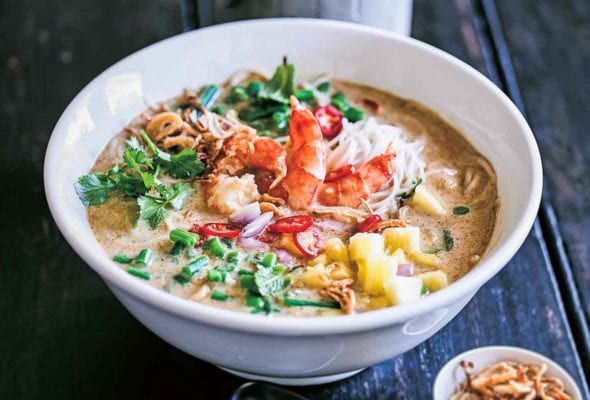 A white bowl of shrimp laksa soup with a small bowl of crispy fried shallots, a spoon, and a container of chopsticks on the side.