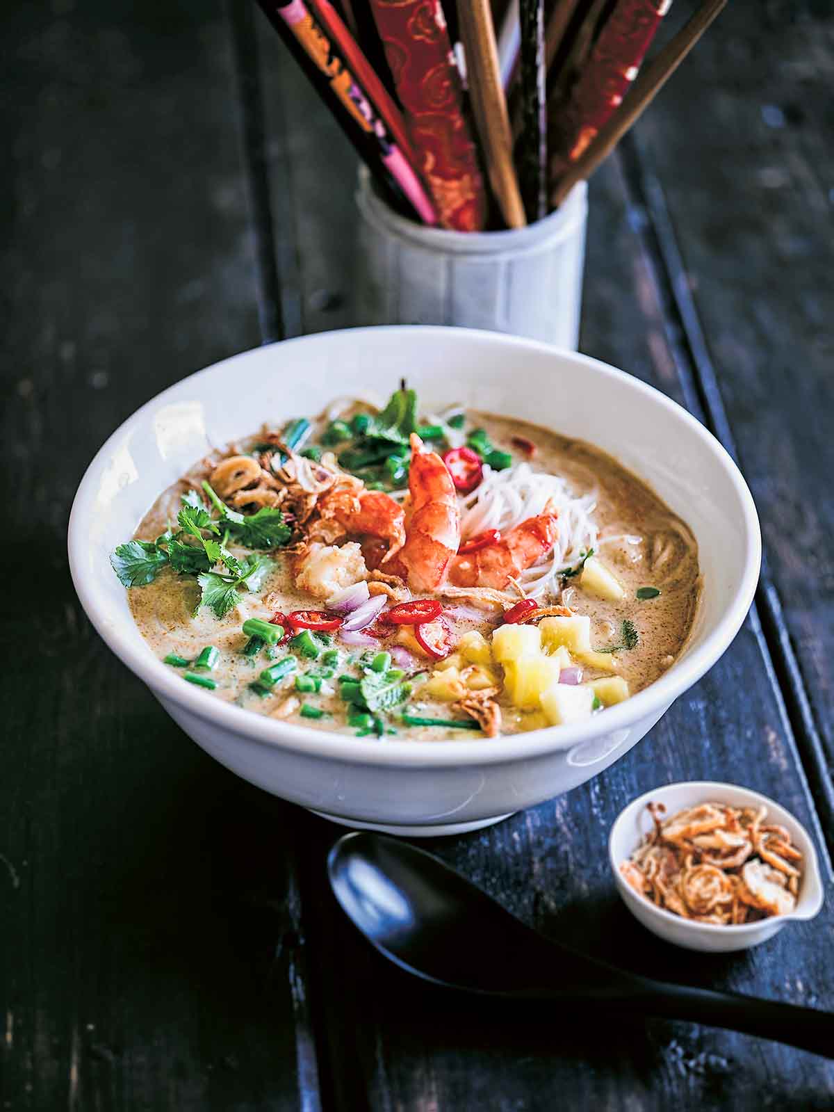 A white bowl of shrimp laksa soup with a small bowl of crispy fried shallots, a spoon, and a container of chopsticks on the side.