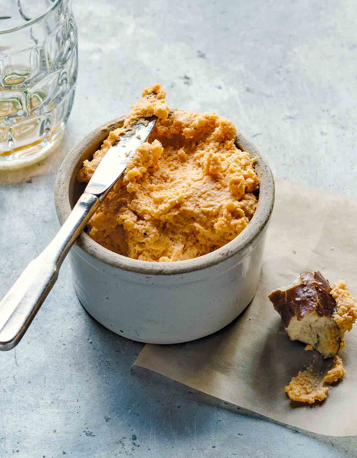 A crock of beer cheese with a knife resting on top, and a mug of beer and piece of pretzel bun on the side.