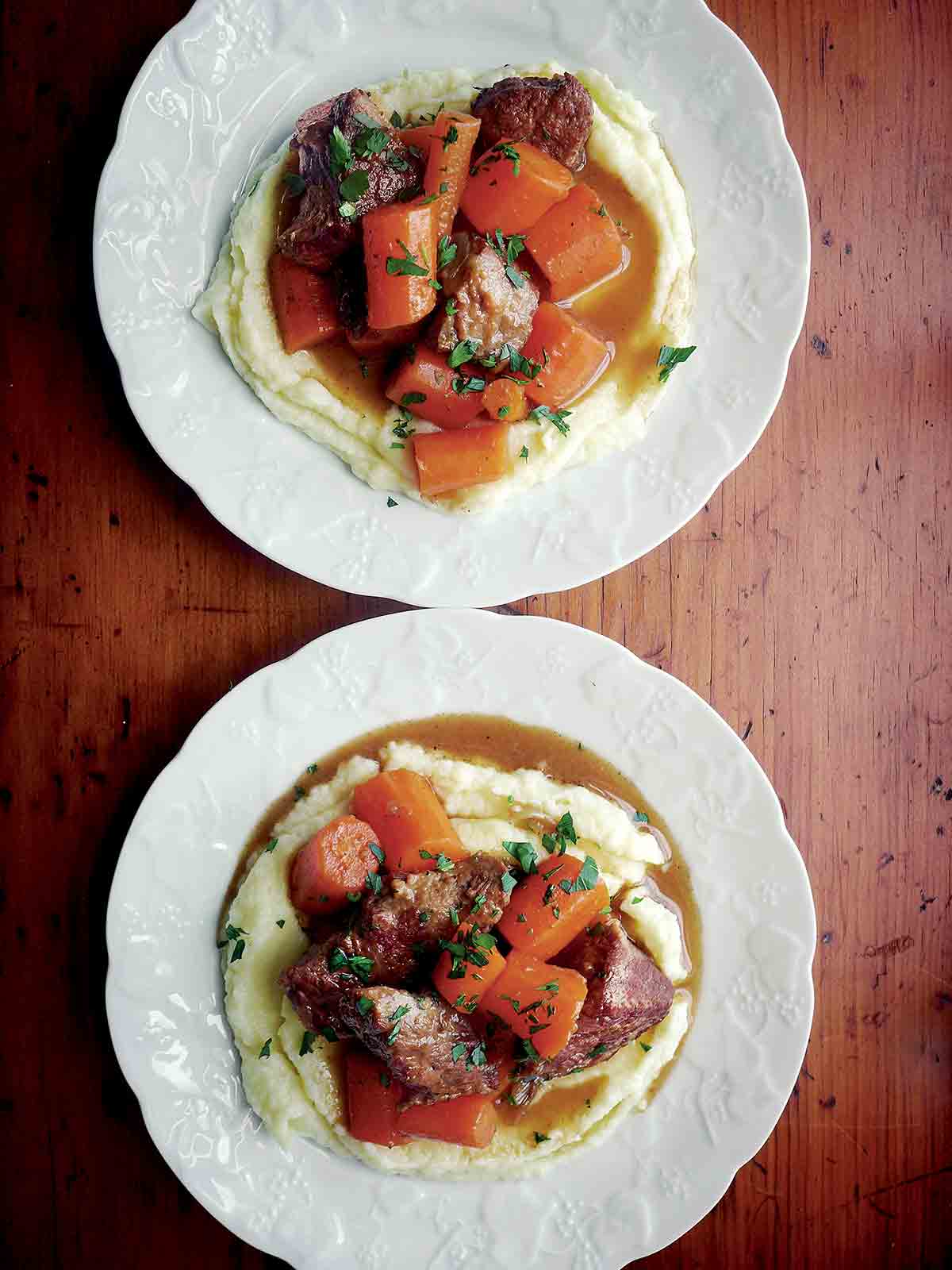 Two white plates of braised beef with carrots on top of mashed potatoes.