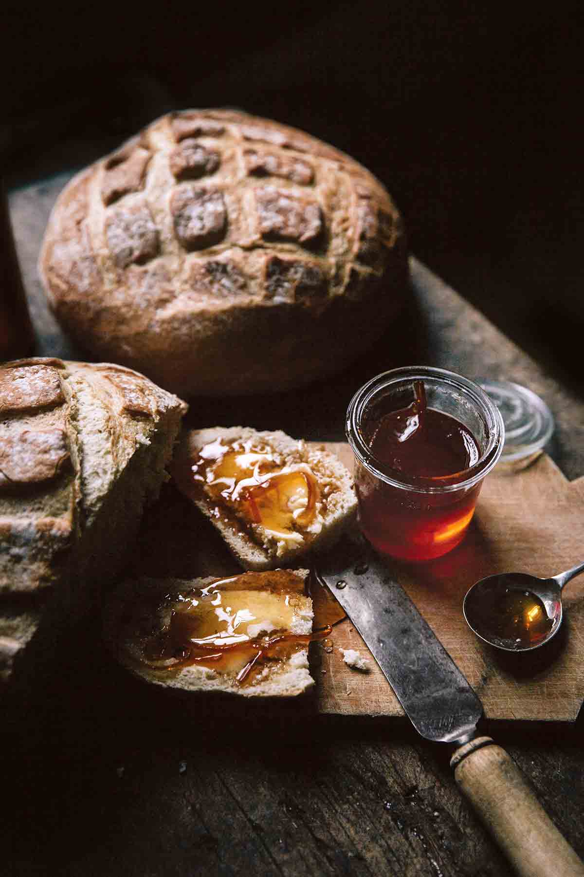 Two loaves of artisan bread and a jar of Campari citrus marmalade, with a slice of bread smeared with the marmalade on a wooden cutting board.