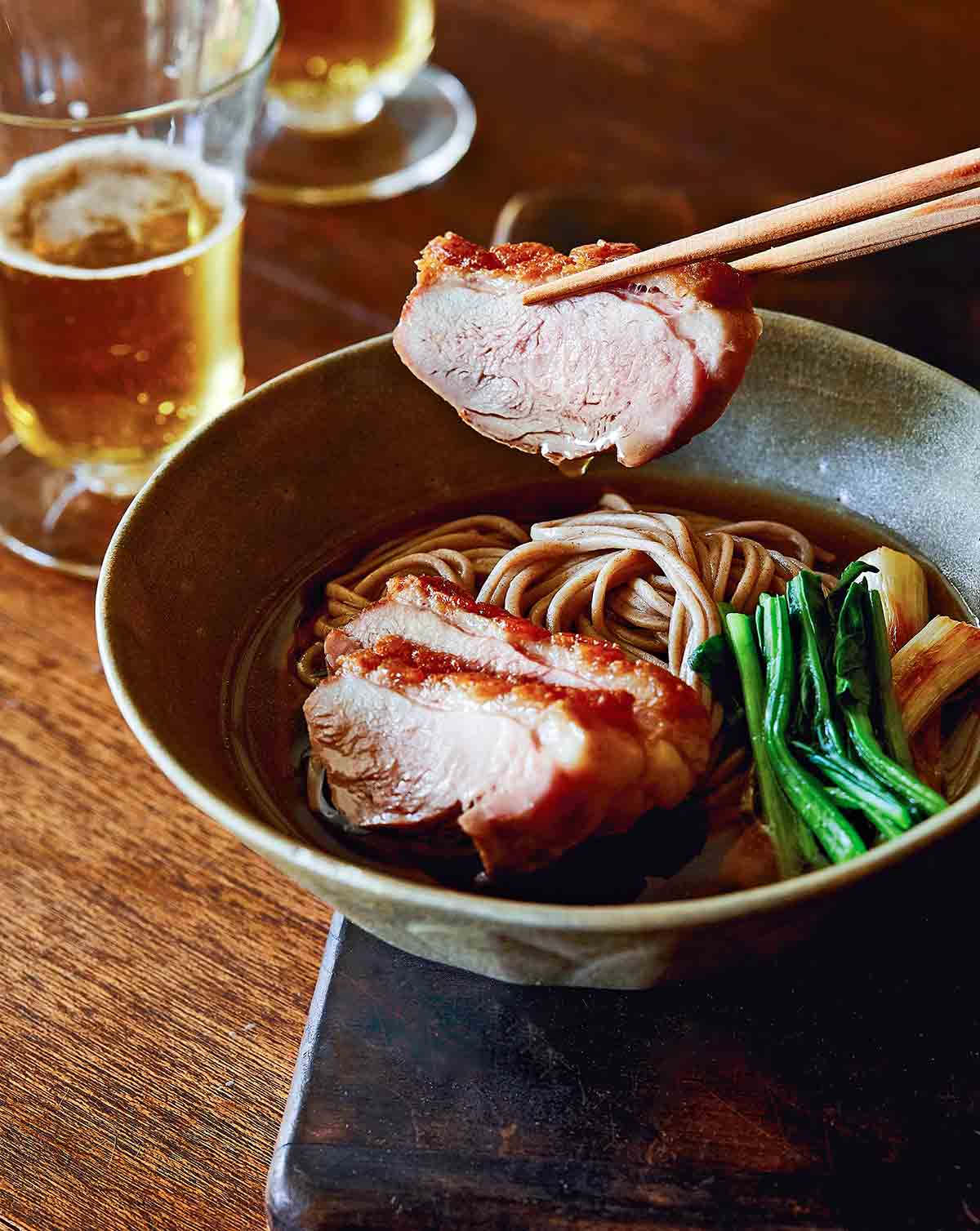 A bowl of duck soba with scallions on a wooden surface with a piece of duck being lifted out with chopsticks.