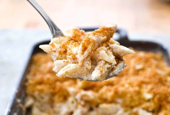 A dark grey rectangular baking dish half filled with four cheese macaroni and cheese and a metal spoon lifting a scoop out.