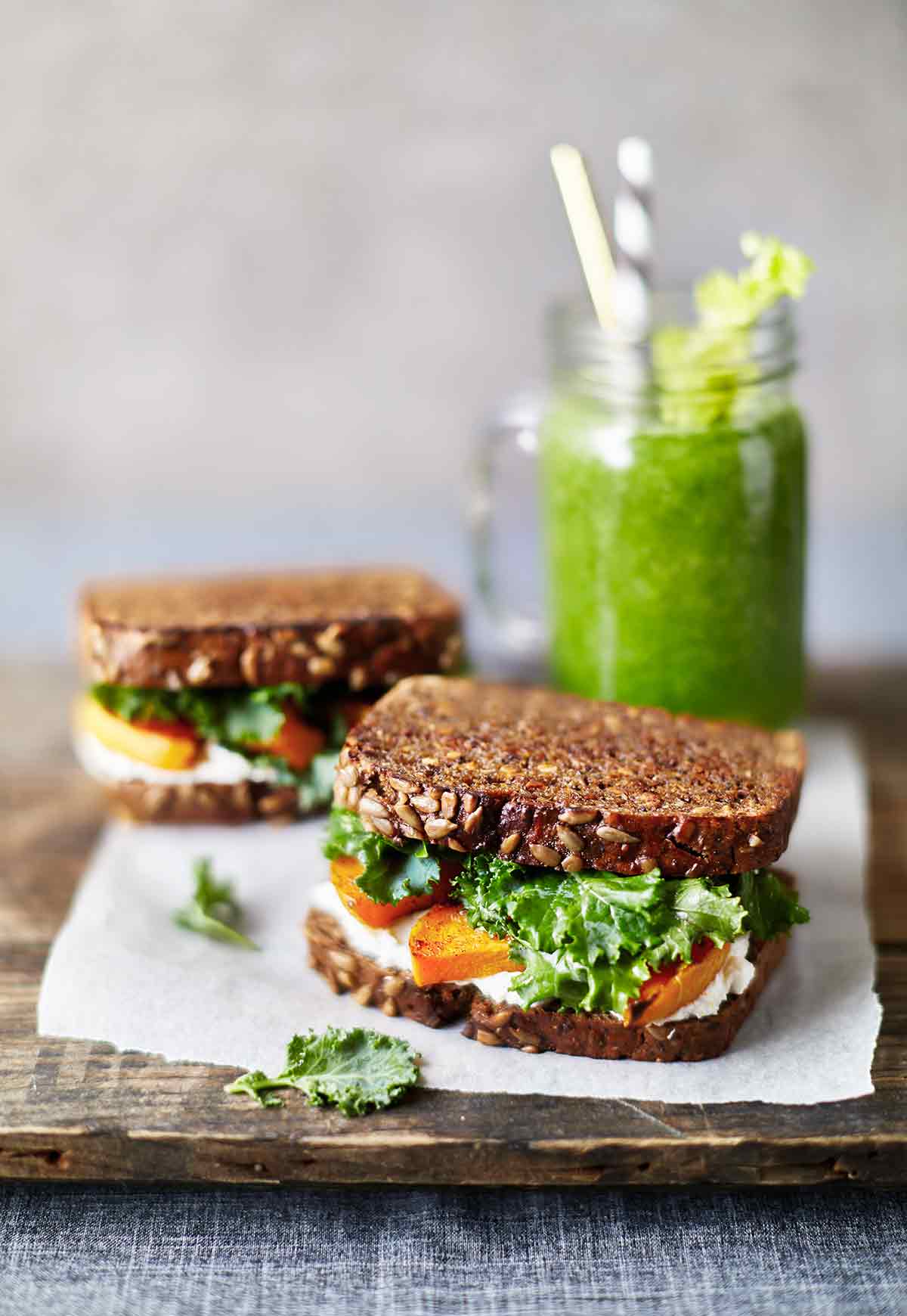 Two grilled cheese with caramelized butternut squash and kale on a pice of white paper with a green smoothie in the background.