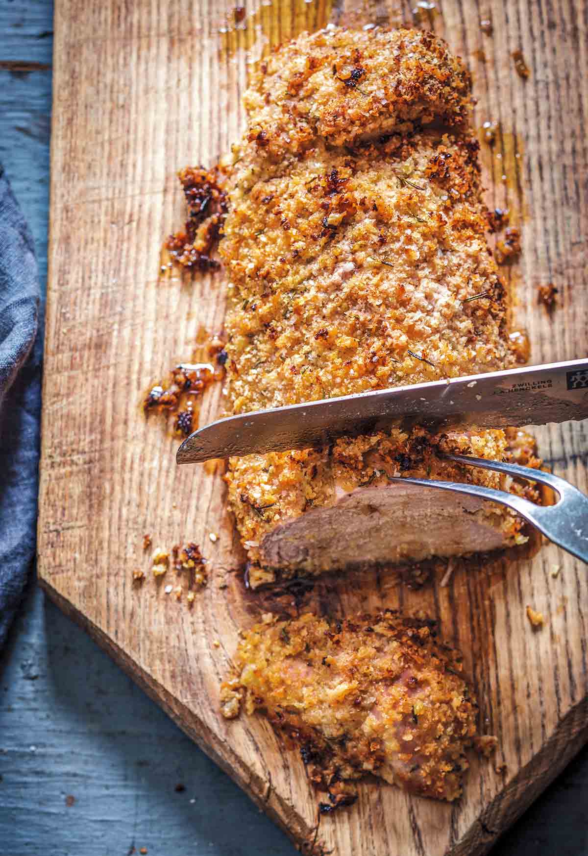 A panko crusted pork tenderloin with rosemary being sliced on a wooden cutting board.