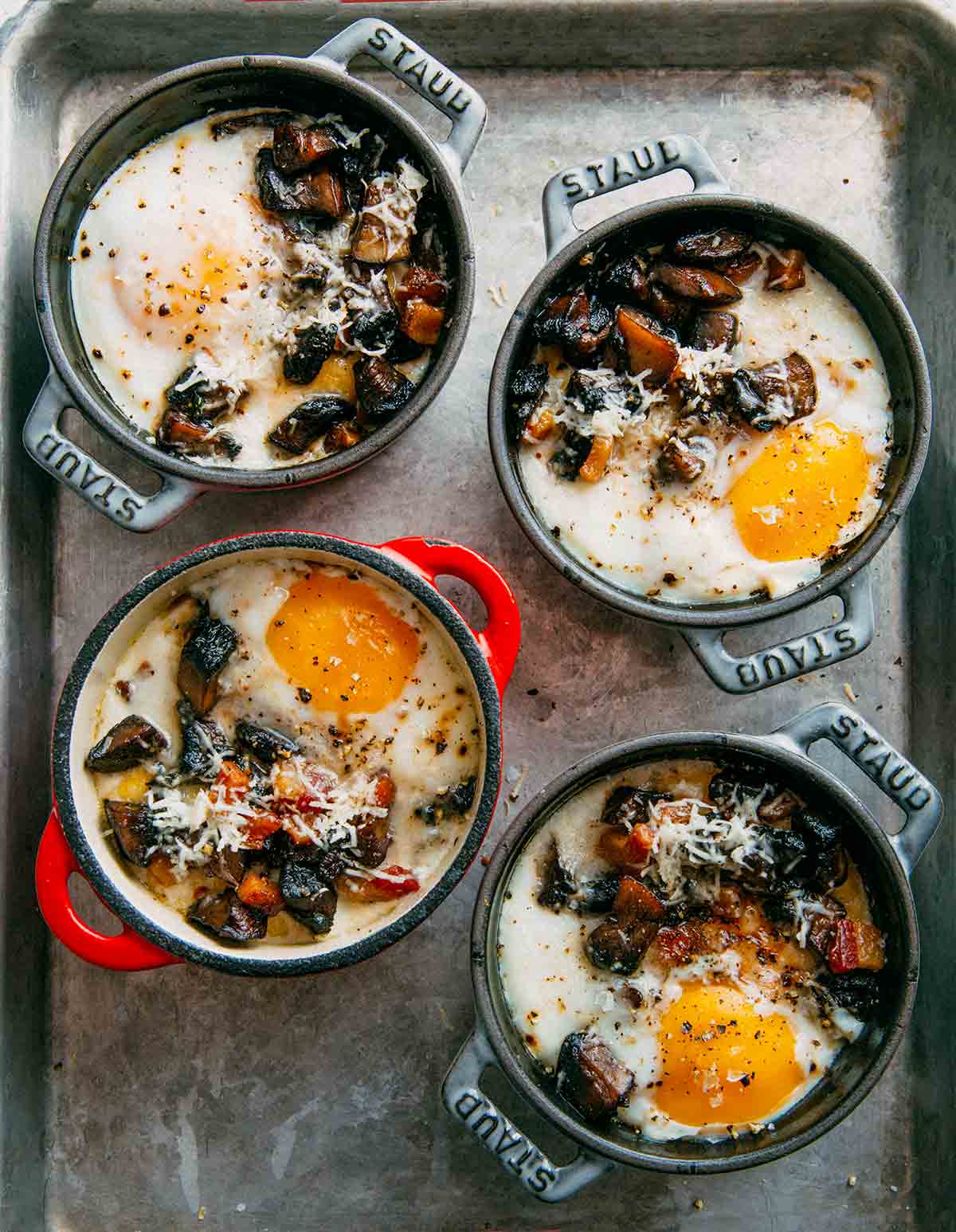 Four individual cocottes filled with baked eggs with polenta and mushrooms on a rimmed baking sheet.