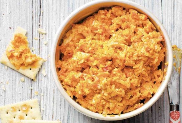A white bowl filled with pimento cheese and some saltine crackers topped with the cheese next to the bowl