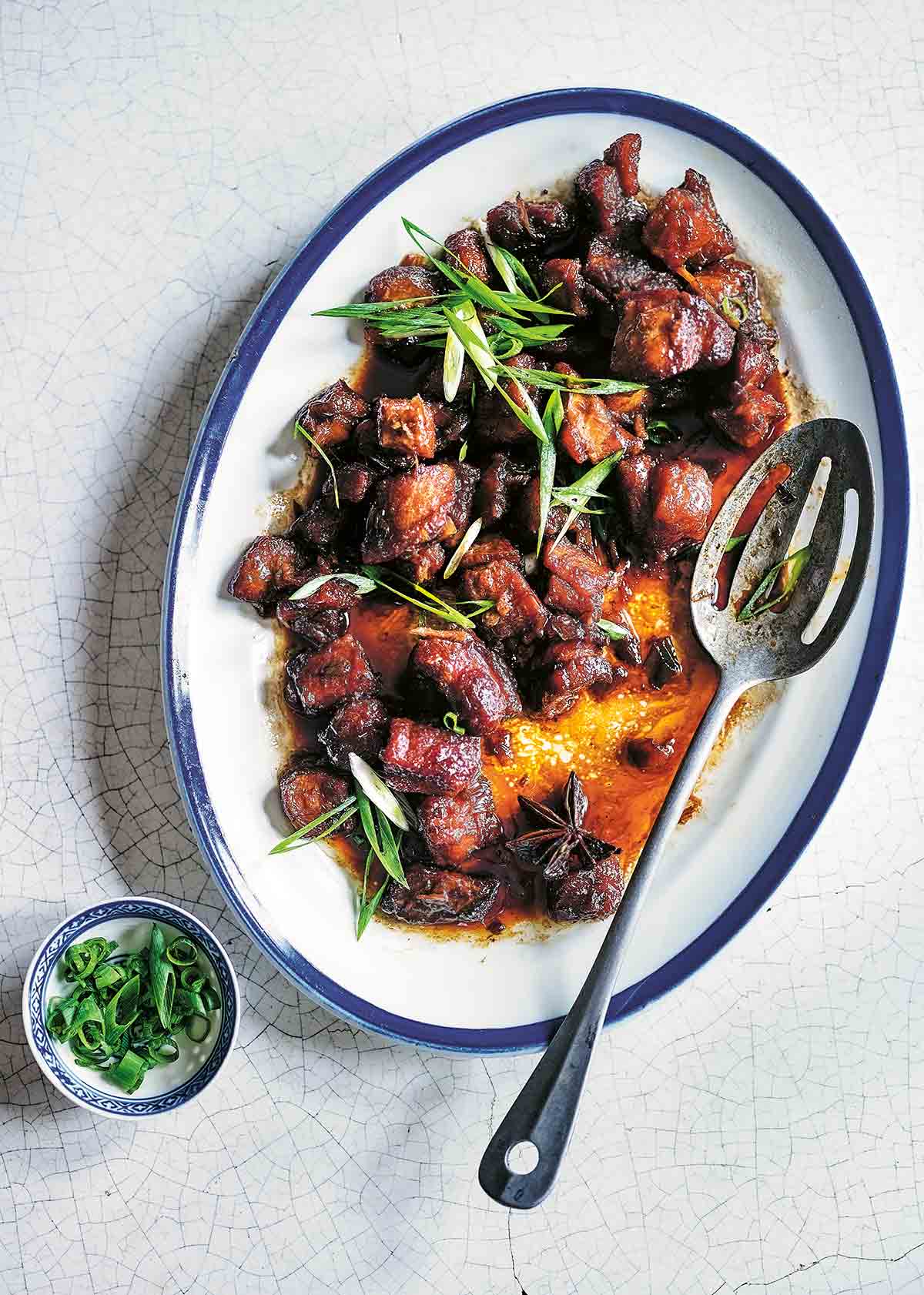 A blue-rimmed oval platter filled with Chairman Mao's red braised pork topped with sliced scallions and a serving spoon resting on the side.