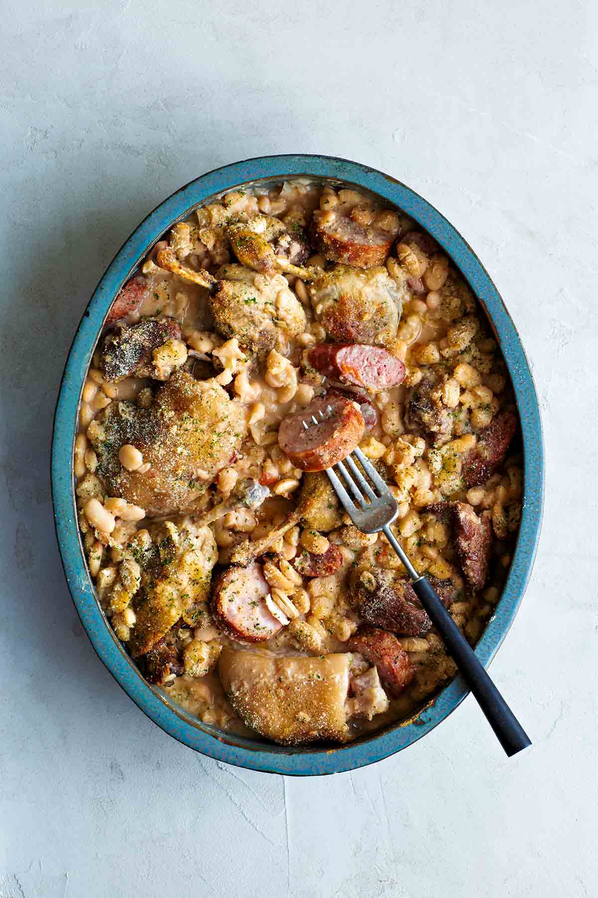 A blue oval ovenproof baking dish filled with classic cassoulet de castelnaudary with a fork resting on top.