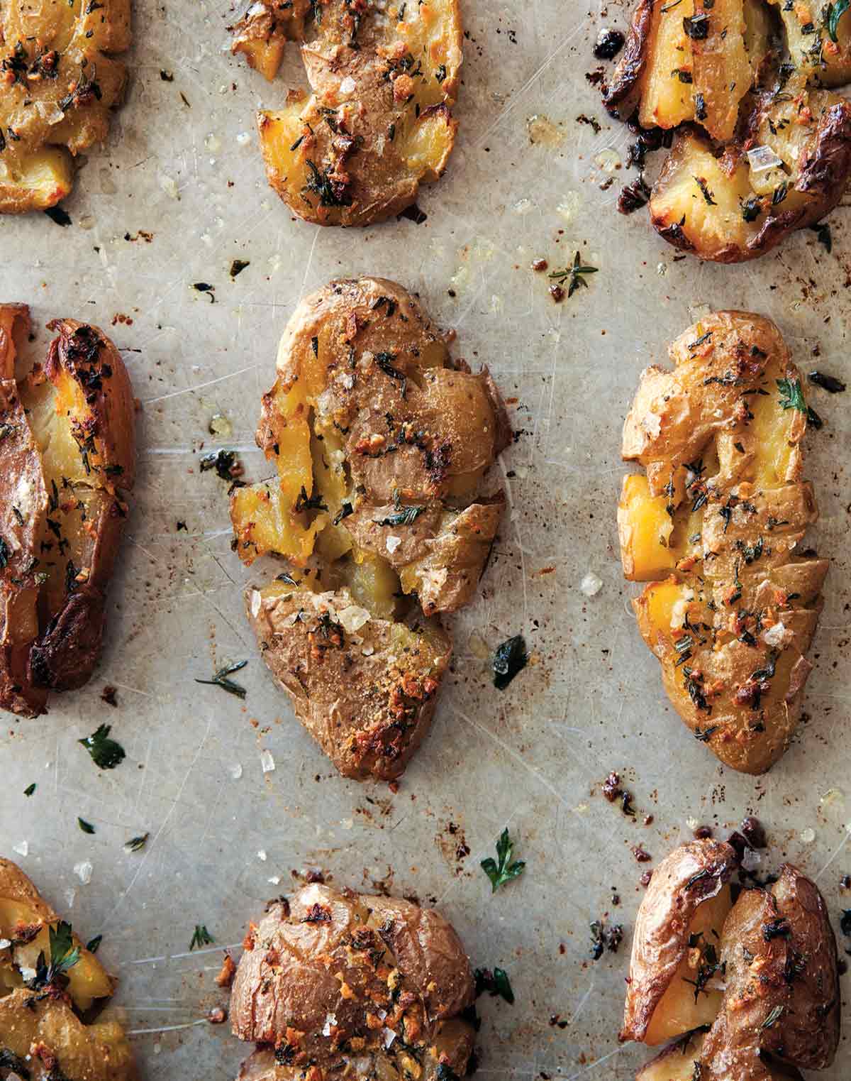 Nine crispy smashed potatoes with herbs on a baking sheet.