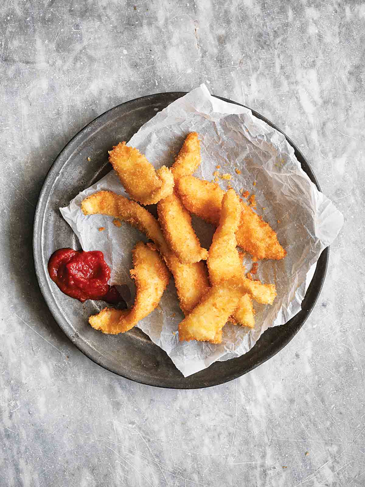 A pile of fancy fish sticks on a piece of parchment with a dollop of ketchup beside them.