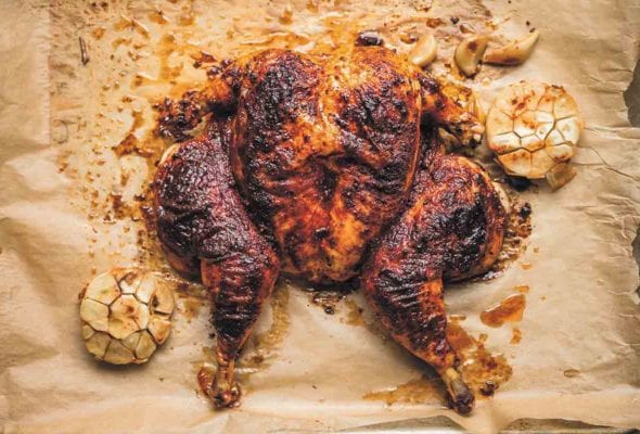 A flat roasted chicken with smoked paprika on a sheet of parchment paper with a halved head of garlic on either side.