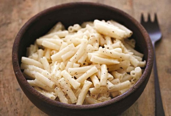 A brown bowl filled with gluten-free macaroni and cheese sprinkled with pepper on a wooden table with a fork resting beside the bowl.